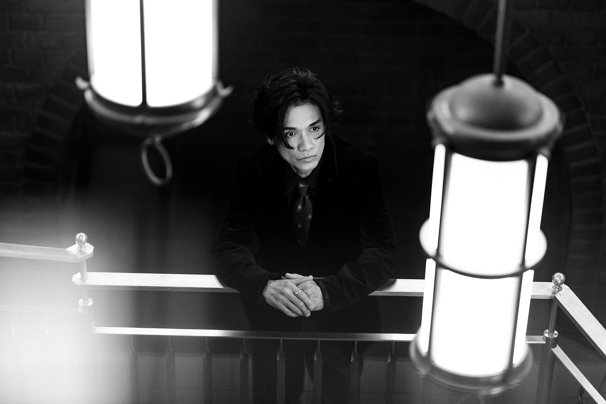 Black-and-white photo of a man in a suit sitting at a desk with hands folded, flanked by bright standing lamps in a dim room.